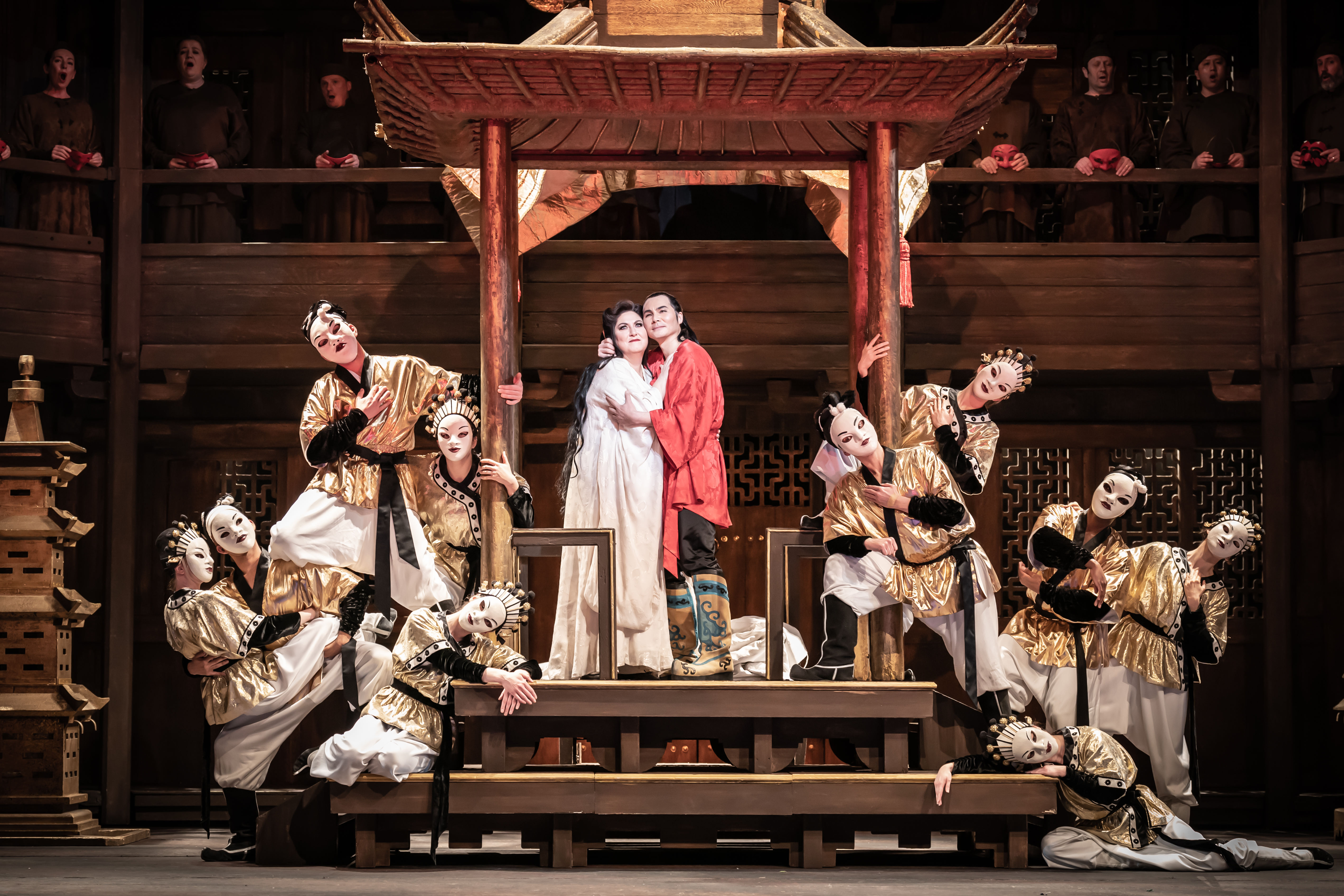 Performers in elaborate costumes and white face makeup pose on a wooden stage, with two central figures embracing under a pagoda-like structure, surrounded by dancers wearing traditional Japanese dance theatre masks. .
