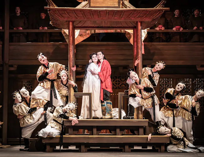 Performers in elaborate costumes and white face makeup pose on a wooden stage, with two central figures embracing under a pagoda-like structure, surrounded by dancers wearing traditional Japanese dance theatre masks. .