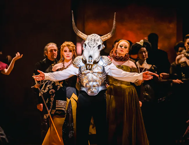 A performer wears a decoratively carved horn skull as a mask and painted steel armour over a white shirt. Their arms are outstretched, and they are holding a sword in their left hand. They are flanked by two women wearing medieval style ballgowns. Behind them are the chorus, all wearing medieval-style clothing.