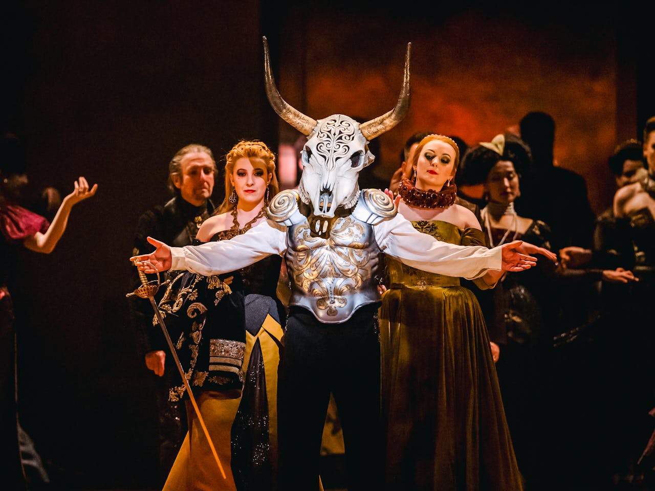 A performer wears a decoratively carved horn skull as a mask and painted steel armour over a white shirt. Their arms are outstretched, and they are holding a sword in their left hand. They are flanked by two women wearing medieval style ballgowns. Behind them are the chorus, all wearing medieval-style clothing. They are performing the Royal Opera production Rigoletto.