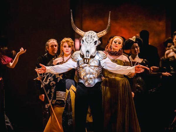 A performer wears a decoratively carved horn skull as a mask and painted steel armour over a white shirt. Their arms are outstretched, and they are holding a sword in their left hand. They are flanked by two women wearing medieval style ballgowns. Behind them are the chorus, all wearing medieval-style clothing. They are performing the Royal Opera production Rigoletto.