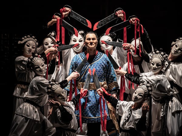 An opera singer wearing a blue Oriental-style tunic with gold banding stands in the centre. Nine performers wearing white Oriental-style tunics and white oriental-style face masks point swords towards the opera singer's head. The swords have red blood stains painted on them and are bound with red ribbon which flows down the performer's arms.