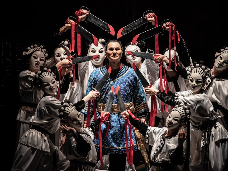 An opera singer wearing a blue Oriental-style tunic with gold banding stands in the centre. Nine performers wearing white Oriental-style tunics and white oriental-style face masks point swords towards the opera singer's head. The swords have red blood stains painted on them and are bound with red ribbon which flows down the performer's arms.
