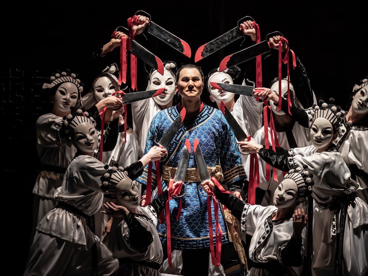 An opera singer wearing a blue Oriental-style tunic with gold banding stands in the centre. Nine performers wearing white Oriental-style tunics and white oriental-style face masks point swords towards the opera singer's head. The swords have red blood stains painted on them and are bound with red ribbon which flows down the performer's arms.