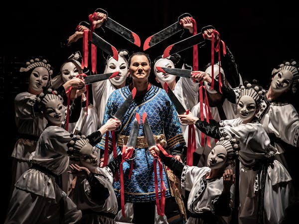 The opera singer Yonghoon Lee is performing as Calaf in The Royal Opera’s Turandot. He is wearing a blue tunic with gold banding and stands in the centre where nine performers surround him. They are wearing white tunics and white face masks point swords towards the opera singer's head. The swords have red drops painted on them and are bound with red ribbon which flows down the performer's arms.