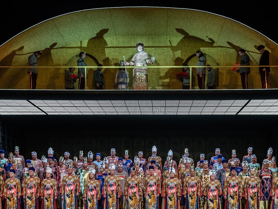 A Chorus of opera singers stand on stage. They are wearing traditional Russian-styled colourful traditional gowns and different styles of Clergy headpieces. Above them, on a platform, stands Bryn Terfel performing as Boris Godunov wearing a gold Russian-style smock and holding a sceptre and orb. There are several performers with him dressed as servents in grey suits. Some are holding red cushions outwards and some are bowing.