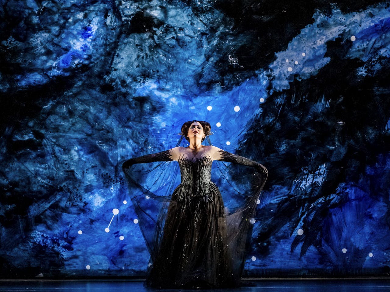 The soprano Tuuli Takala performs as the Queen of the Night in The Magic Flute. She stands in the centre of the stage, her arms spread outwards with the mesh was her long black ballgown by her wrists. The stage behind her is painted in blue and black brushstrokes with white dots making up the constellation of stars.