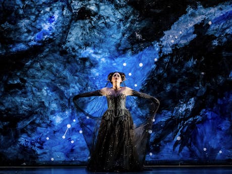 Tuuli Takala is performing as The Queen of The Night in The Royal Opera’s production of The Magic Flute. She stands in the centre of the stage with their arms spread outwards. The mesh from her long black ballgown draped out from her wrists. The stage behind her is set in blue and black brushstrokes with white dots making up the constellation of stars.