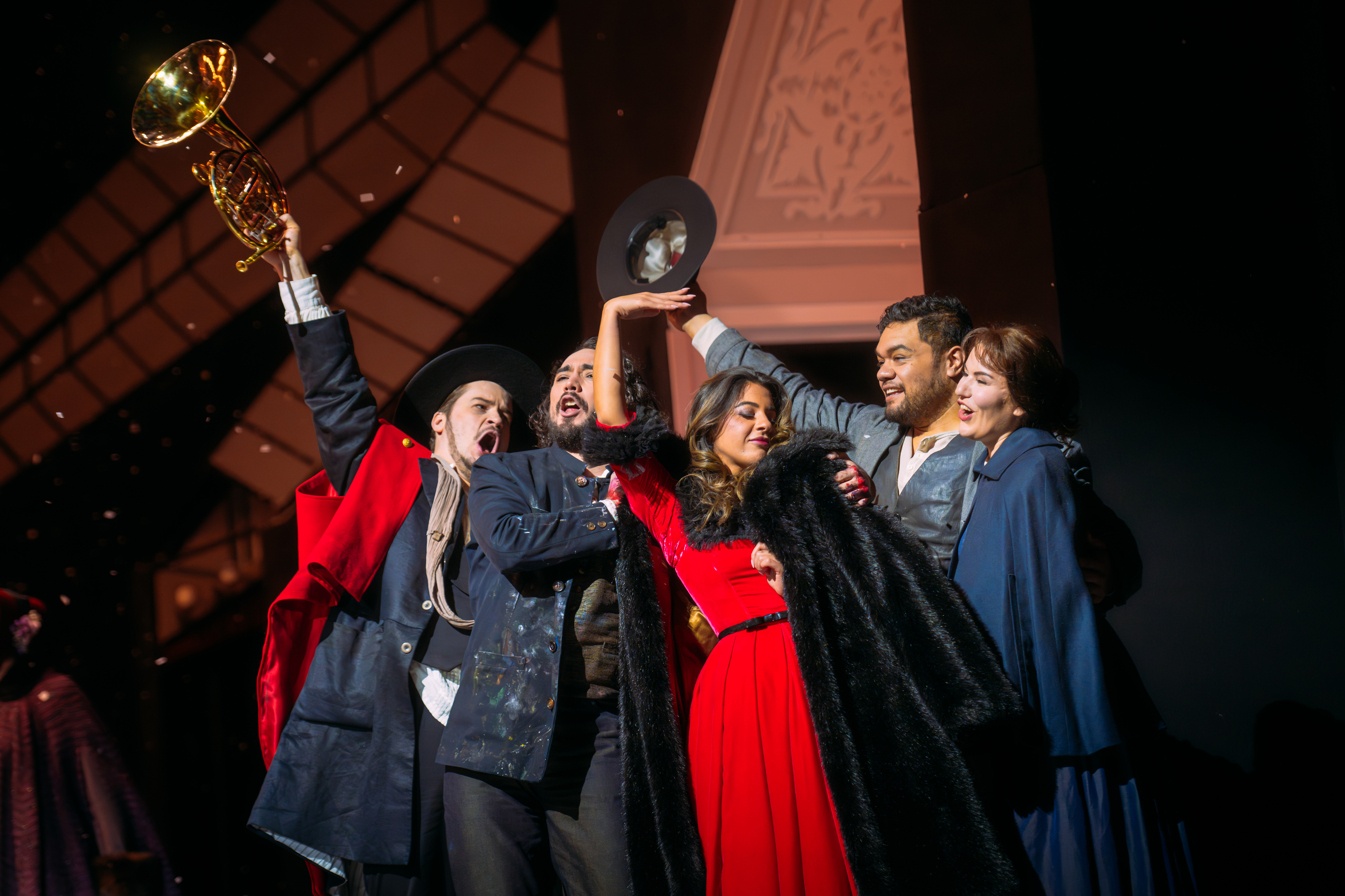Five people stand huddled together. One man on the outside of the huddle holds out a French horn into the air. A woman in a plush red dress with black fur trim holds a black fur coat close to her. Another person holds up their hat into the air.  
