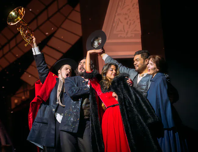 Five people stand huddled together. One man on the outside of the huddle holds out a French horn into the air. A woman in a plush red dress with black fur trim holds a black fur coat close to her. Another person holds up their hat into the air. (3 of 4)
