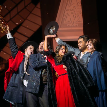 Five people stand huddled together. One man on the outside of the huddle holds out a French horn into the air. A woman in a plush red dress with black fur trim holds a black fur coat close to her. Another person holds up their hat into the air.