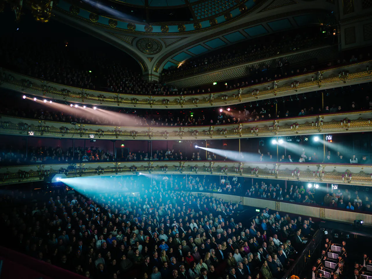 A view across the Main Stage auditorium of The Royal Opera House during a busy performance. The auditorium is dark but several bright white light cones stream across the audience from different parts of the theatre.