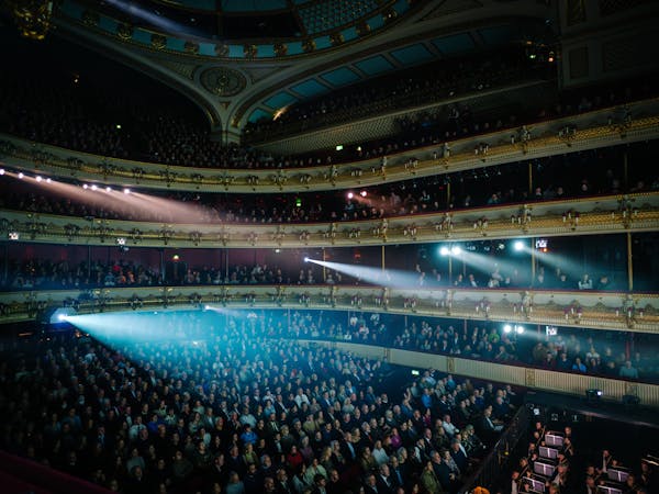 A view across the Main Stage auditorium of The Royal Opera House during a busy performance. The auditorium is dark but several bright white light cones stream across the audience from different parts of the theatre.