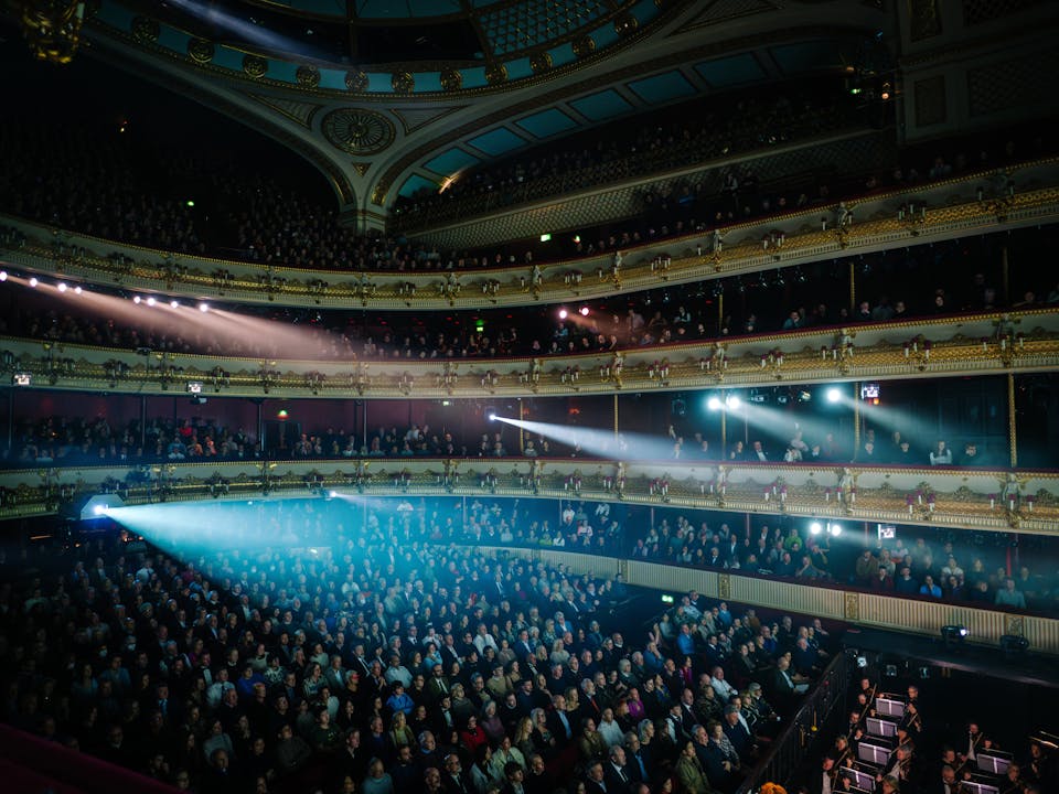 A view across the Main Stage auditorium of The Royal Opera House during a busy performance. The auditorium is dark but several bright white light cones stream across the audience from different parts of the theatre.
