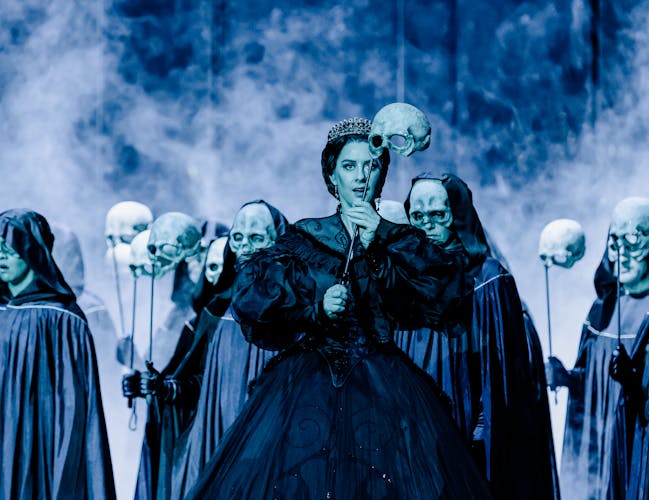 A performer stands on a dark purple lit stage wearing a black Victorian-style gown and a tiara. She holds above her head the top part of a human skull on a cane. Behind her, performers wearing hooded black capes also hold up skulls on canes and some are wearing masks that look like the skulls being held up.