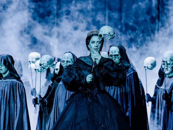 The opera singer Malin Byström stands on a dark purple lit stage wearing a black Victorian-style gown and a tiara at the Royal Opera House in the production of The Sicilian Vespers. She holds above her head the top part of a human skull on a cane. Behind her, performers wearing hooded black capes also hold up skulls on canes and some are wearing masks that look like the skulls being held up.