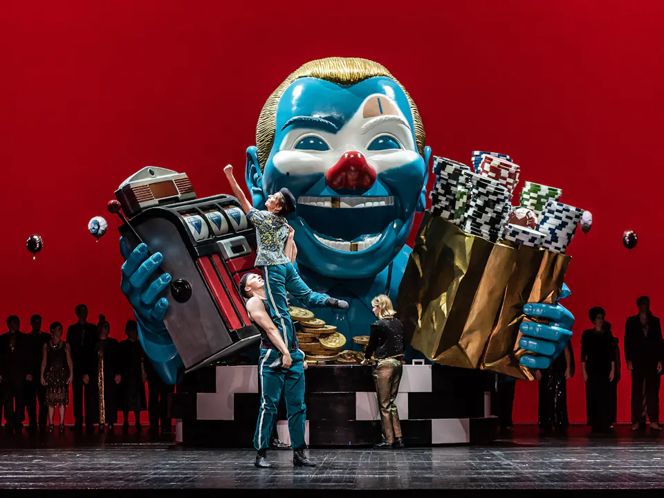 In the centre of the stage stands a large sculpture of a clown’s torso. They have a blue face, gold hair, a red nose and a few gold teeth and hold up a slot machine in one arm and a gold bag filled with arcade chips in the other. A performer wearing blue striped tracksuit bottoms holds up another performer wearing the same clothes. Behind them stands a performer wearing gold trousers. The stage is backlit with eerie red lighting which shows the outline of a line of performers.
