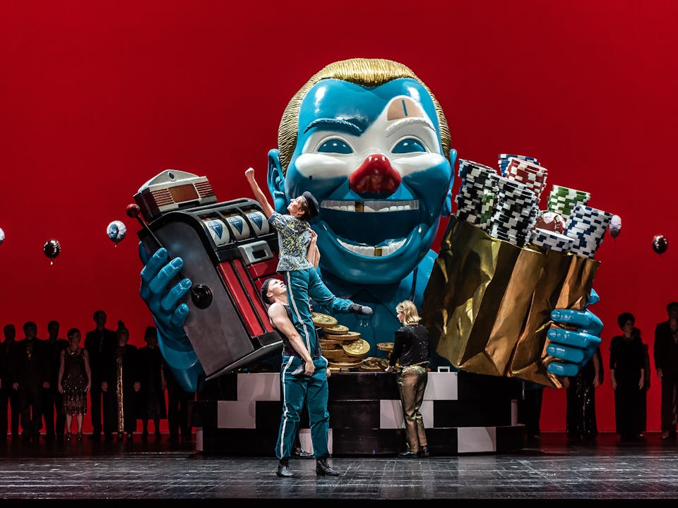 In the center of the stage stands a large sculpture of a clown’s torso. They have a blue face, gold hair, a red nose and a few gold teeth and hold up a slot machine in one arm and a gold bag filled with arcade chips in the other. A performer wearing blue striped tracksuit bottoms holds up another performer wearing the same clothes. Behind them stands a performer wearing gold trousers. The stage is backlit with eerie red lighting which shows the outline of a line of performers. There are several helium balloons with the clown's face scattered on stage. They are performing the opera Samson and Delilah.