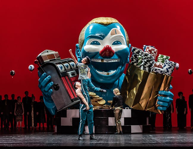 In the centre of the stage stands a large sculpture of a clown’s torso. They have a blue face, gold hair, a red nose and a few gold teeth and hold up a slot machine in one arm and a gold bag filled with arcade chips in the other. A performer wearing blue striped tracksuit bottoms holds up another performer wearing the same clothes. Behind them stands a performer wearing gold trousers. The stage is backlit with eerie red lighting which shows the outline of a line of performers.