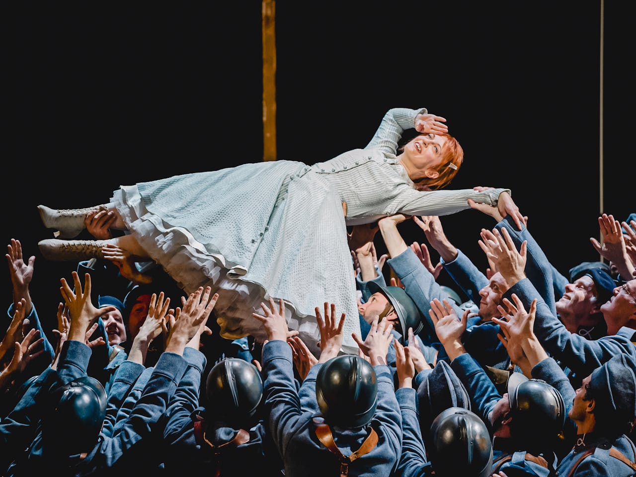 The Soprano Patrizia Ciofi performs in The Royal Opera’s production of La fille du regiment. She has red hair and is wearing a light green striped gown with white lace-up boots. She is being held up in the air, on her back, by dozens of performers dressed as soldiers wearing army uniform, berets or black helmets. She holds her right hand up to her head in a salute.