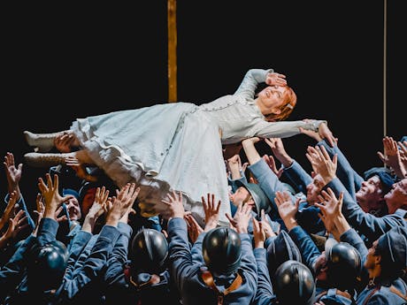 The Soprano Patrizia Ciofi performs in The Royal Opera’s production of La fille du regiment. She has red hair and is wearing a light green striped gown with white lace-up boots. She is being held up in the air, on her back, by dozens of performers dressed as soldiers wearing army uniform, berets or black helmets. She holds her right hand up to her head in a salute.