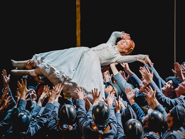 The Soprano Patrizia Ciofi performs in The Royal Opera’s production of La fille du regiment. She has red hair and is wearing a light green striped gown with white lace-up boots. She is being held up in the air, on her back, by dozens of performers dressed as soldiers wearing army uniform, berets or black helmets. She holds her right hand up to her head in a salute.