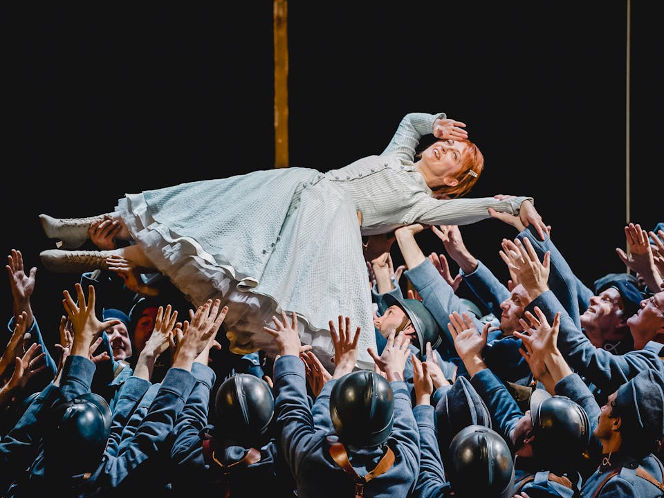 The Soprano Patrizia Ciofi performs in The Royal Opera’s production of La fille du regiment. She has red hair and is wearing a light green striped gown with white lace-up boots. She is being held up in the air, on her back, by dozens of performers dressed as soldiers wearing army uniform, berets or black helmets. She holds her right hand up to her head in a salute.