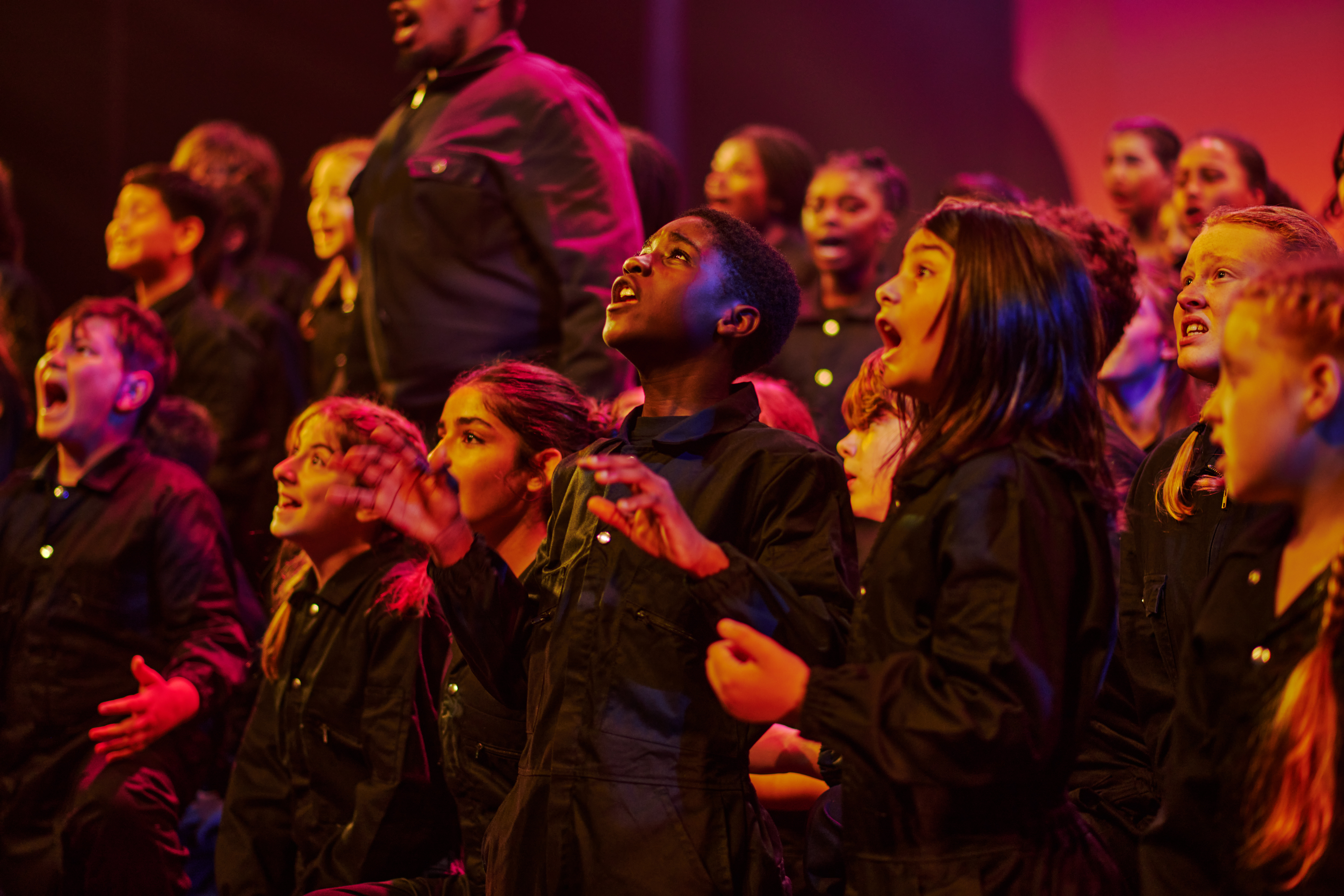 A group of children dressed in black sing on a stage. The lighting is tinged pink and purple.