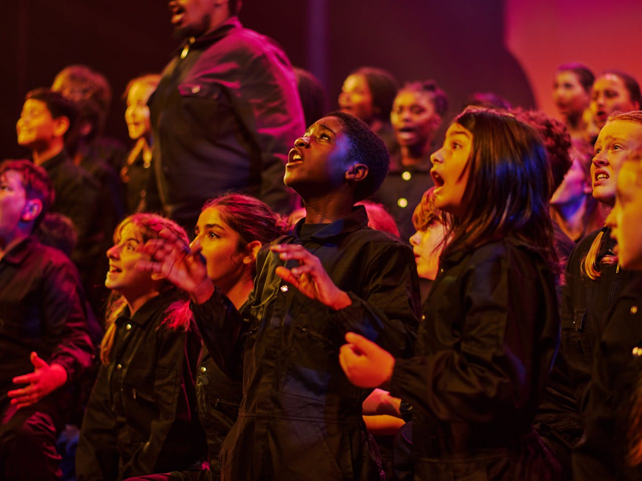 A group of children dressed in black sing on a stage. The lighting is tinged pink and purple.