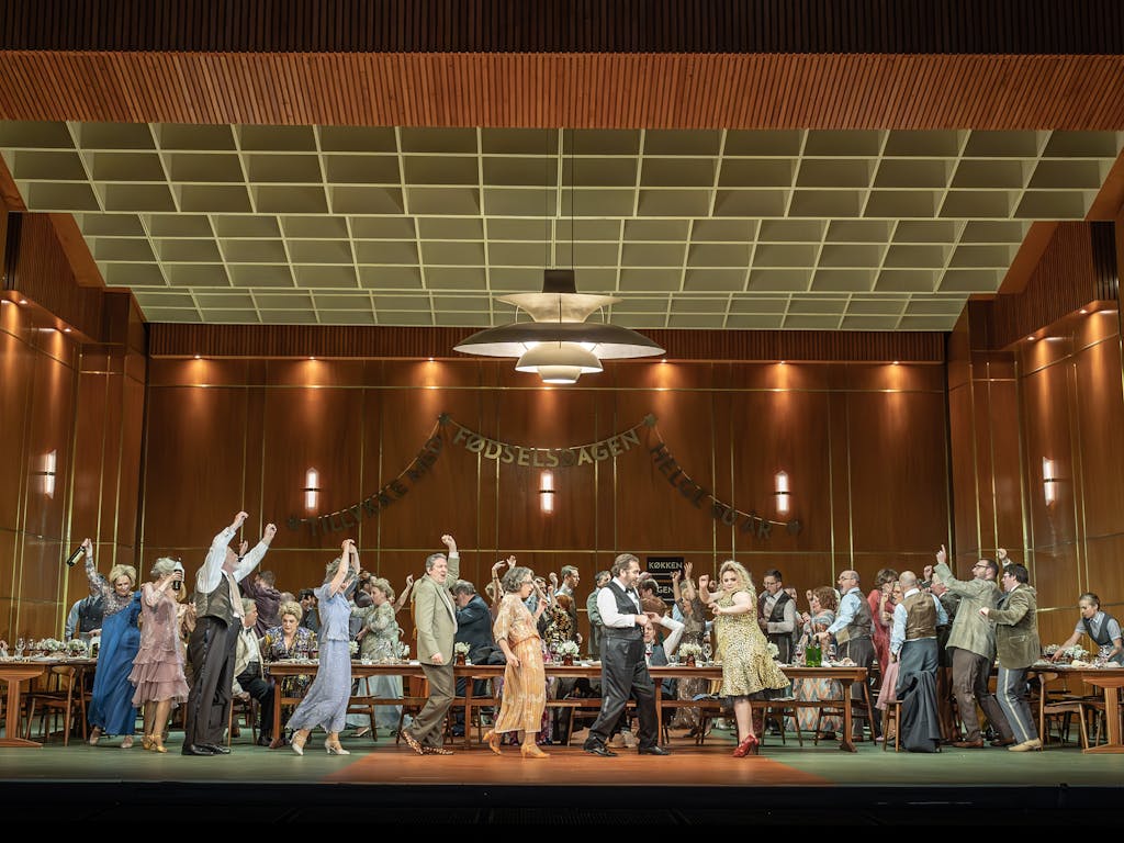 The stage is set with brown wood panel walls. A birthday banner is hung on the back wall. In the centre, there is a long wooden table laid with drinks. A large party of people are dancing around the table, some have their arms and drinks raised in the air.