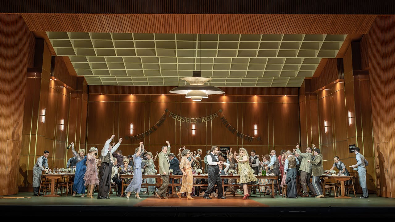 The stage is set with brown wood panel walls. A birthday banner is hung on the back wall. In the centre, there is a long wooden table laid with drinks. A large party of people are dancing around the table, some have their arms and drinks raised in the air.
