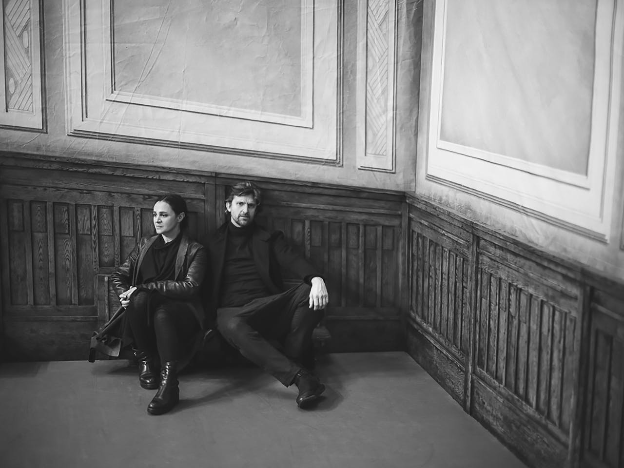 Two people wearing all black clothing sit on the floor of a wooden paneled room – one wears a long leather jacket. The top half of the room is light coloured panel and the bottom half is dark coloured paneling. The image is black and white and is from a press shoot for Sol León and Paul Lightfoot’s production So are We at The Royal Ballet.