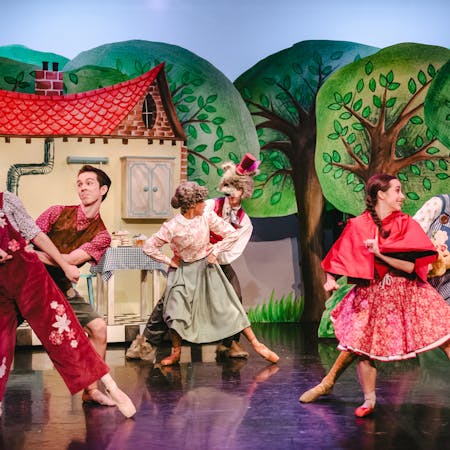 A ballet dancer wearing red pointe ballet slippers, a red and white flower-patterned corseted dress and a red cape over her shoulder's dances in front of a dancer wearing blue and white striped overalls and a gold-coloured furry bear mask and hat. In the centre, another dancer wearing a green skirt, blouse and a grey curly wig performs a similar move in front of a dancer wearing a white shirt, red waistcoat and a grey furry wolf mask with a red top hat. On the other side two dancers join them. The stage is set with tall hand drawn green trees and a little house with a red roof that overlap one another so they appear 3D.