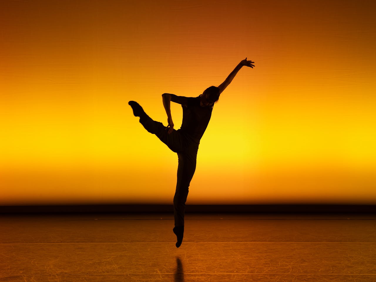 A ballet dancer performs in the centre of a golden-lit stage making it look like a sunset. Only the outline of their body is visible making them appear to be in a shadow. They are contorted into a Y-shape, with their right foot en pointe and their left leg high in the air.