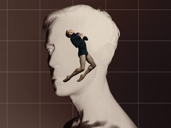 The ballet Edward Watson wears a black long-sleeve rollneck leotard and jumps into the air. His legs are bent, and his toes point towards the floor. At this chest, his hands are clutched together. Behind him, the stage is set with an image of a brown grid background and inverted photograph of a man with short chair and striking features head and neck which makes the image appear entirely white – almost as though it is a sculpture. He is performing in The Royal Ballet’s production of A Single Man.