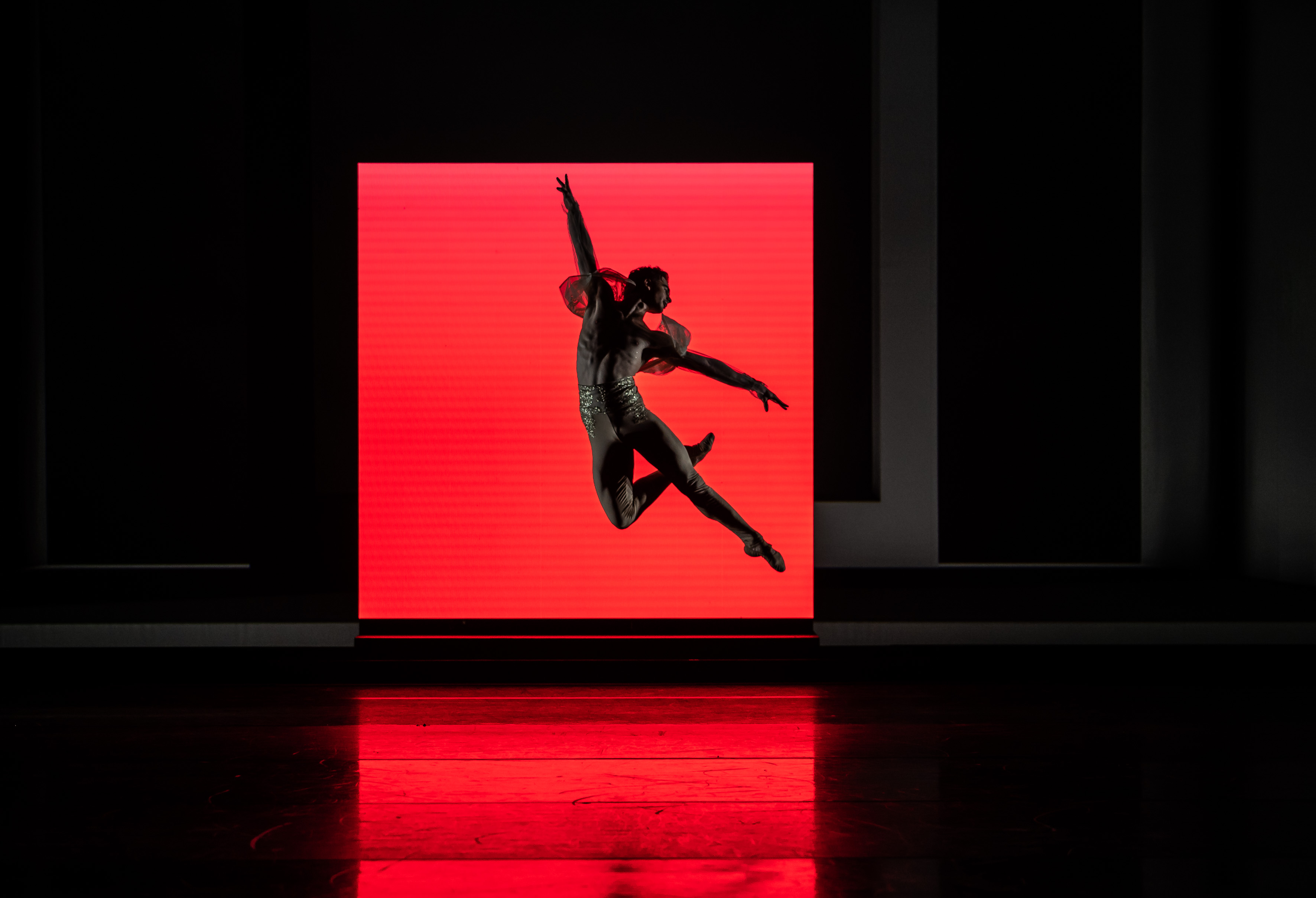 A ballet dancer wearing nude leggings decorated with gold sequins at the waistband launches themselves into the air – their right knee is bent behind their left leg which points out straight, their arms are stretched outwards creating a T shape and their head is turned towards their shoulders. They are jumping in front of a red brightly lit square which creates a dark shadow on the whole stage.    