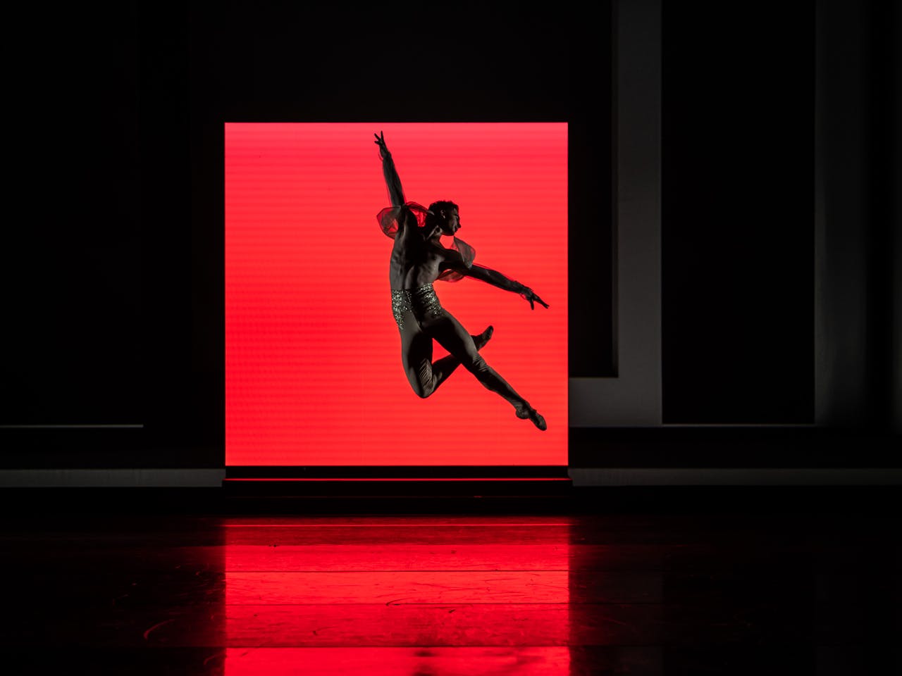 A ballet dancer wearing nude leggings decorated with gold sequins at the waistband launches themselves into the air – their right knee is bent behind their left leg which points out straight, their arms are stretched outwards creating a T shape and their head is turned towards their shoulders. They are jumping in front of a red brightly lit square which creates a dark shadow on the whole stage.