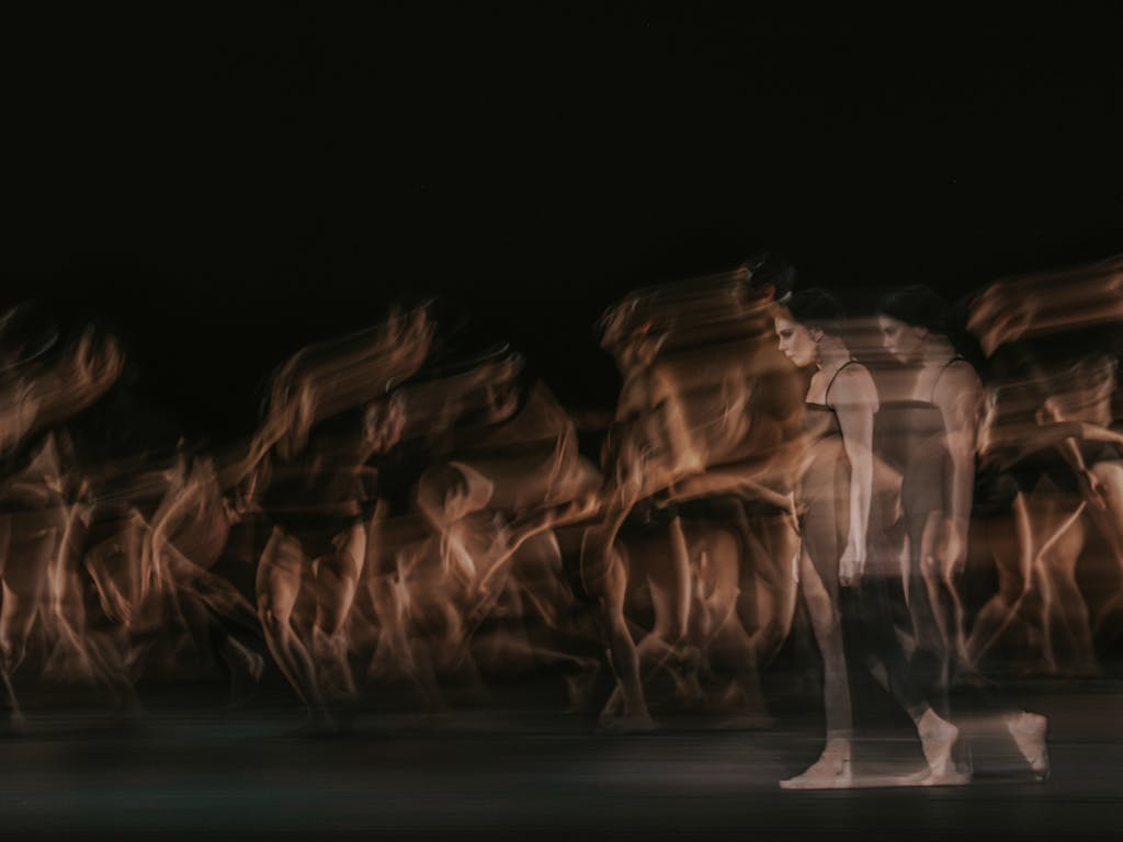 The ballet dancer Natalia Osipova walks on stage in a black dress and bare feet. She appears transparent and blurry, her image doubled behind her as though in movement. Behind her, multiple ballet dancers are blurry as though in movement. They are performing in The Royal Ballet’s production of Woolf Works.