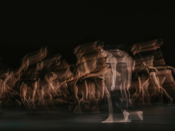 The ballet dancer Natalia Osipova walks on stage in a black dress and bare feet. She appears transparent and blurry, her image doubled behind her as though in movement. Behind her, multiple ballet dancers are blurry as though in movement. They are performing in The Royal Ballet’s production of Woolf Works.