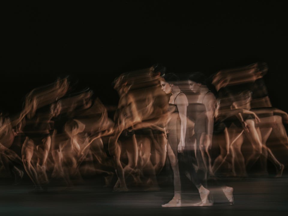 The ballet dancer Natalia Osipova walks on stage in a black dress and bare feet. She appears transparent and blurry, her image doubled behind her as though in movement. Behind her, multiple ballet dancers are blurry as though in movement. They are performing in The Royal Ballet’s production of Woolf Works.