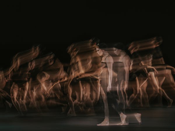 The ballet dancer Natalia Osipova walks on stage in a black dress and bare feet. She appears transparent and blurry, her image doubled behind her as though in movement. Behind her, multiple ballet dancers are blurry as though in movement. They are performing in The Royal Ballet’s production of Woolf Works.