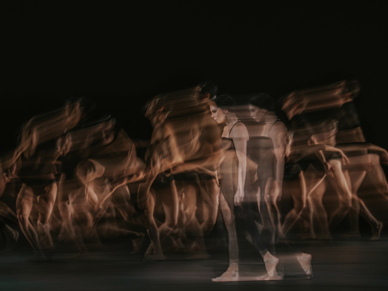 The ballet dancer Natalia Osipova walks on stage in a black dress and bare feet. She appears transparent and blurry, her image doubled behind her as though in movement. Behind her, multiple ballet dancers are blurry as though in movement. They are performing in The Royal Ballet’s production of Woolf Works.