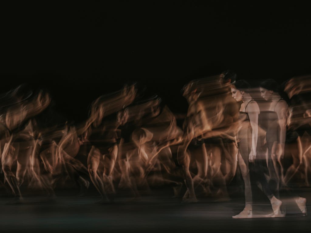 The ballet dancer Natalia Osipova walks on stage in a black dress and bare feet. She appears transparent and blurry, her image doubled behind her as though in movement. Behind her, multiple ballet dancers are blurry as though in movement. They are performing in The Royal Ballet’s production of Woolf Works.