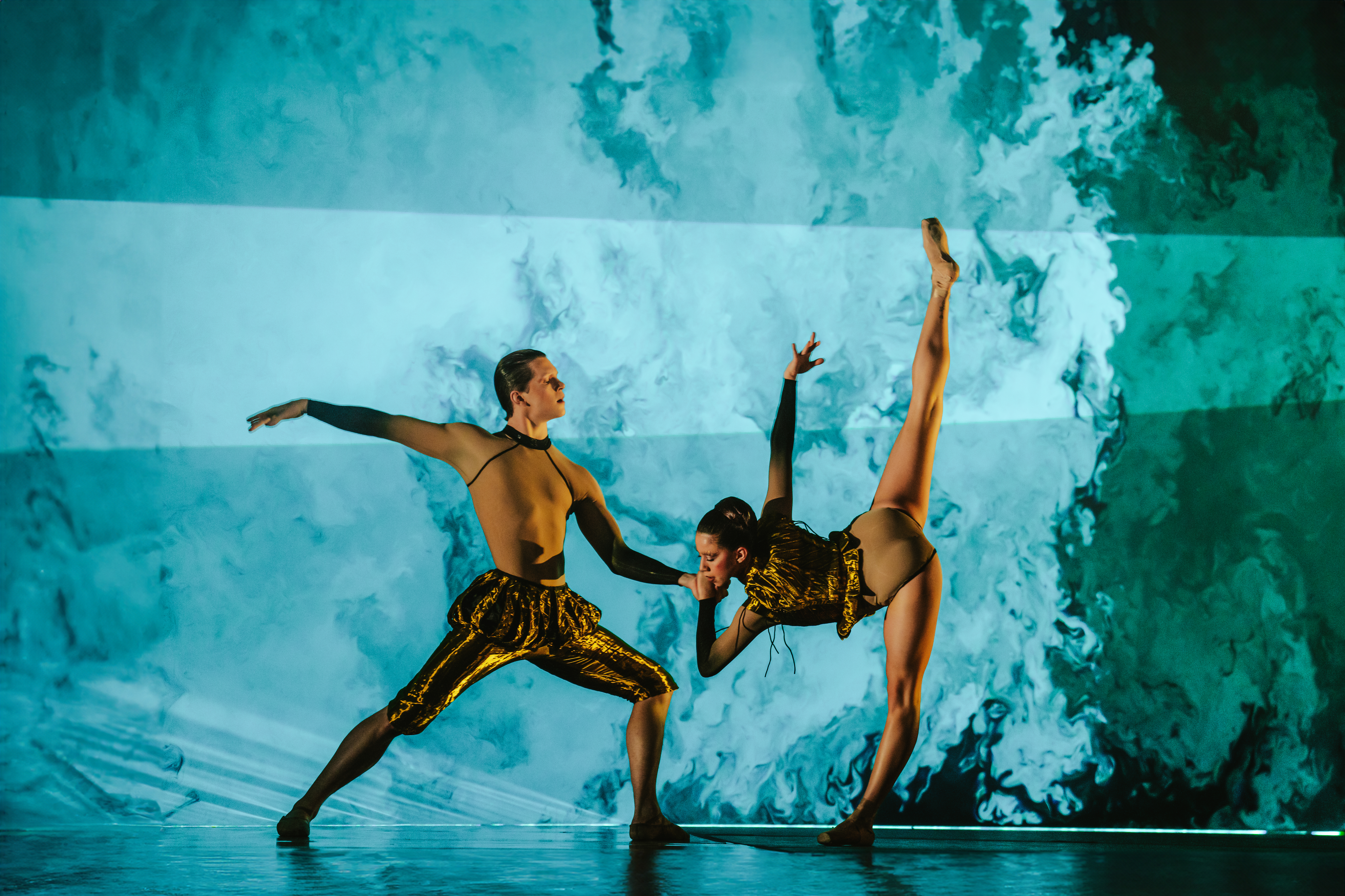 The Royal Ballet Dancers Harris Bell and Leticia Dias stand on stage set with blue and dark green projections that appear like brushstrokes to look like the sea as they perform in The Royal Ballet’s production of Wayne McGregor’s Woolf Works. One dancer wearing gold trousers with frilled gathering at the waist stands in a squat position with their left leg bent and their right leg stretched out behind them and their arms are stretched outwards at their shoulders. The other dancer wearing a gold leotard stands leaning forwards on their left leg, their right leg stretched out vertically to create a long straight shape with their legs. They hold the other dancers hand and their right arm follows the same vertical patter as their legs.   