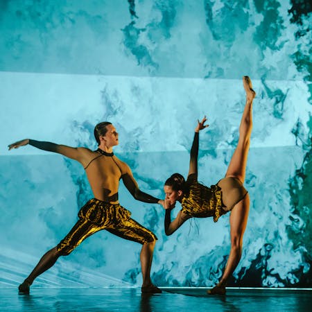 The Royal Ballet Dancers Harris Bell and Leticia Dias stand on stage set with blue and dark green projections that appear like brushstrokes to look like the sea as they perform in The Royal Ballet’s production of Wayne McGregor’s Woolf Works. One dancer wearing gold trousers with frilled gathering at the waist stands in a squat position with their left leg bent and their right leg stretched out behind them and their arms are stretched outwards at their shoulders. The other dancer wearing a gold leotard stands leaning forwards on their left leg, their right leg stretched out vertically to create a long straight shape with their legs. They hold the other dancers hand and their right arm follows the same vertical patter as their legs.
