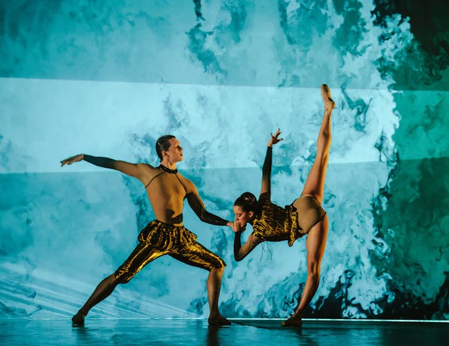 A ballet dancer wearing gold metallic trousers and a sheer top stands with their left leg bent forwards and their right leg stretched straight behind them. They hold onto the hands of another dancer wearing a gold metallic leotard who is standing on their left left while their right one stretched straight above into the air. The stage is set with washed blue and green background and lighting.