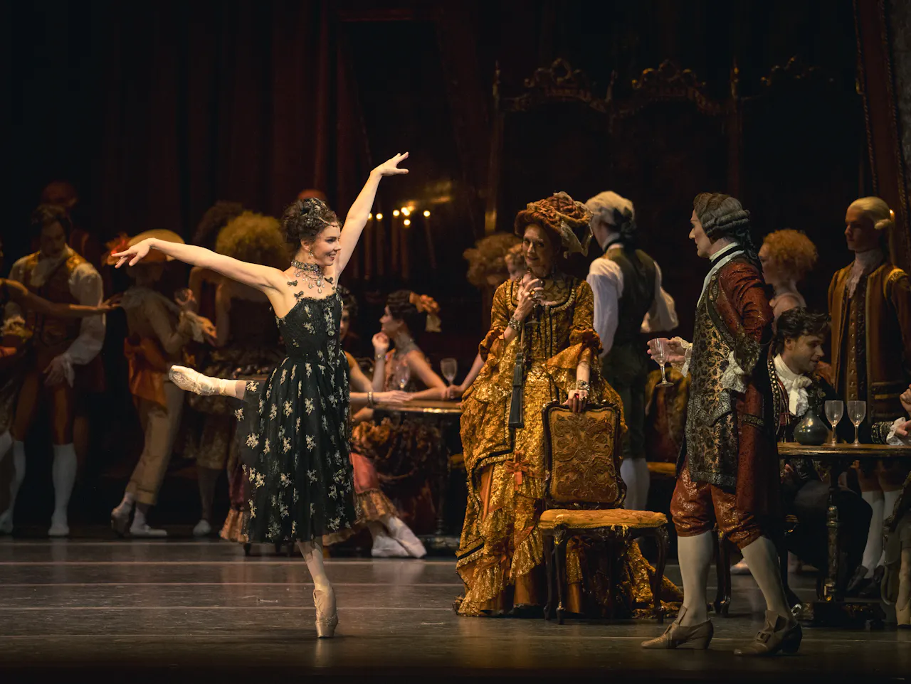 A male dancer wearing green tights and a brown shirt is holding a woman wearing a brown dress. She is leaning back with her leg outstretched across him, as he lunges near her.