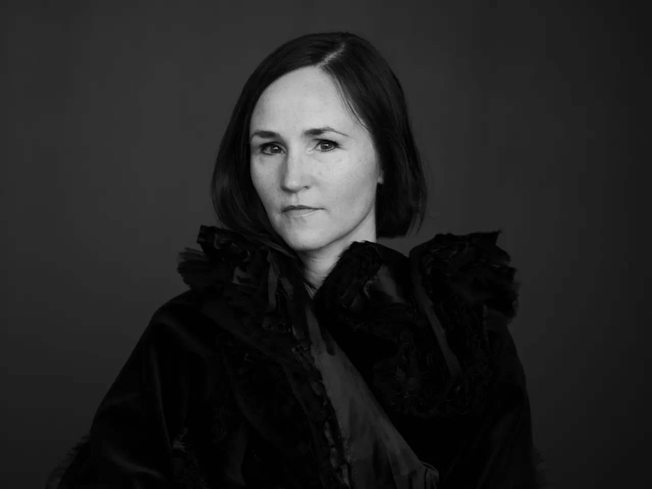 A black and white headshot of the composer Anna Thorvaldsdottir/ She wears a black caped coat and sits against a dark backdrop. On her finger is a large black ring.