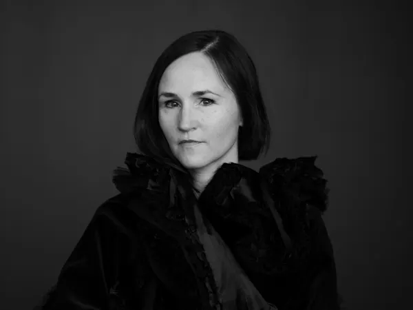 A black and white headshot of the composer Anna Thorvaldsdottir/ She wears a black caped coat and sits against a dark backdrop. On her finger is a large black ring.