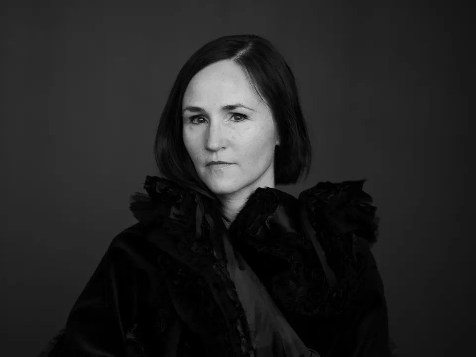 A black and white headshot of the composer Anna Thorvaldsdottir/ She wears a black caped coat and sits against a dark backdrop. On her finger is a large black ring.